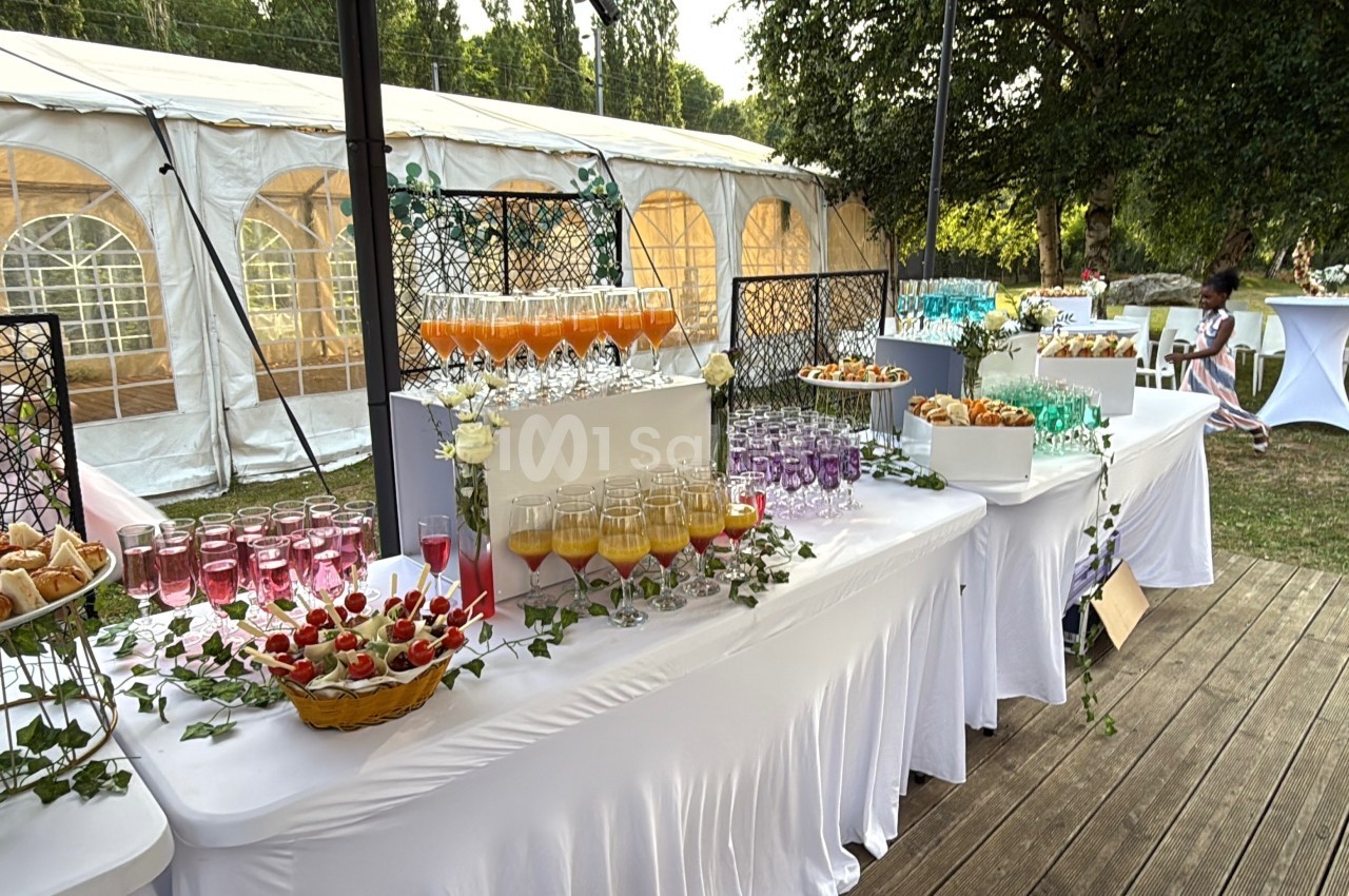 Buffet en plein air avec boissons colorées, verrines et pâtisseries, installé près d'une tente blanche et d'un jardin.