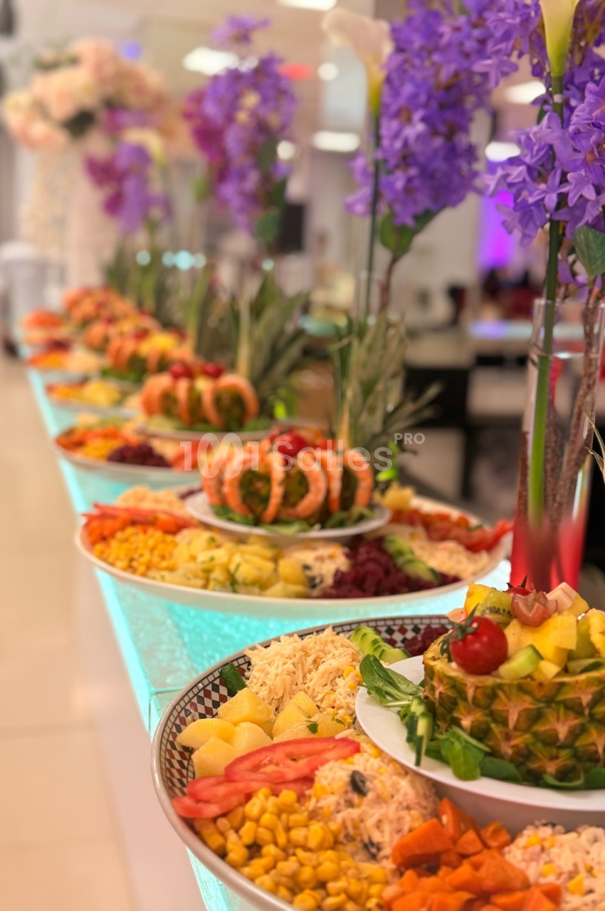 Buffet coloré avec divers plats, fruits et légumes décoratifs, disposés sur une table éclairée, ornée de fleurs violettes.