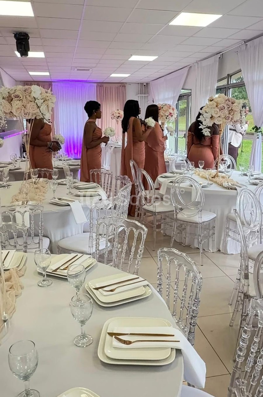 Des femmes en robes roses discutent dans une salle de réception décorée de tables élégantes et de fleurs blanches.