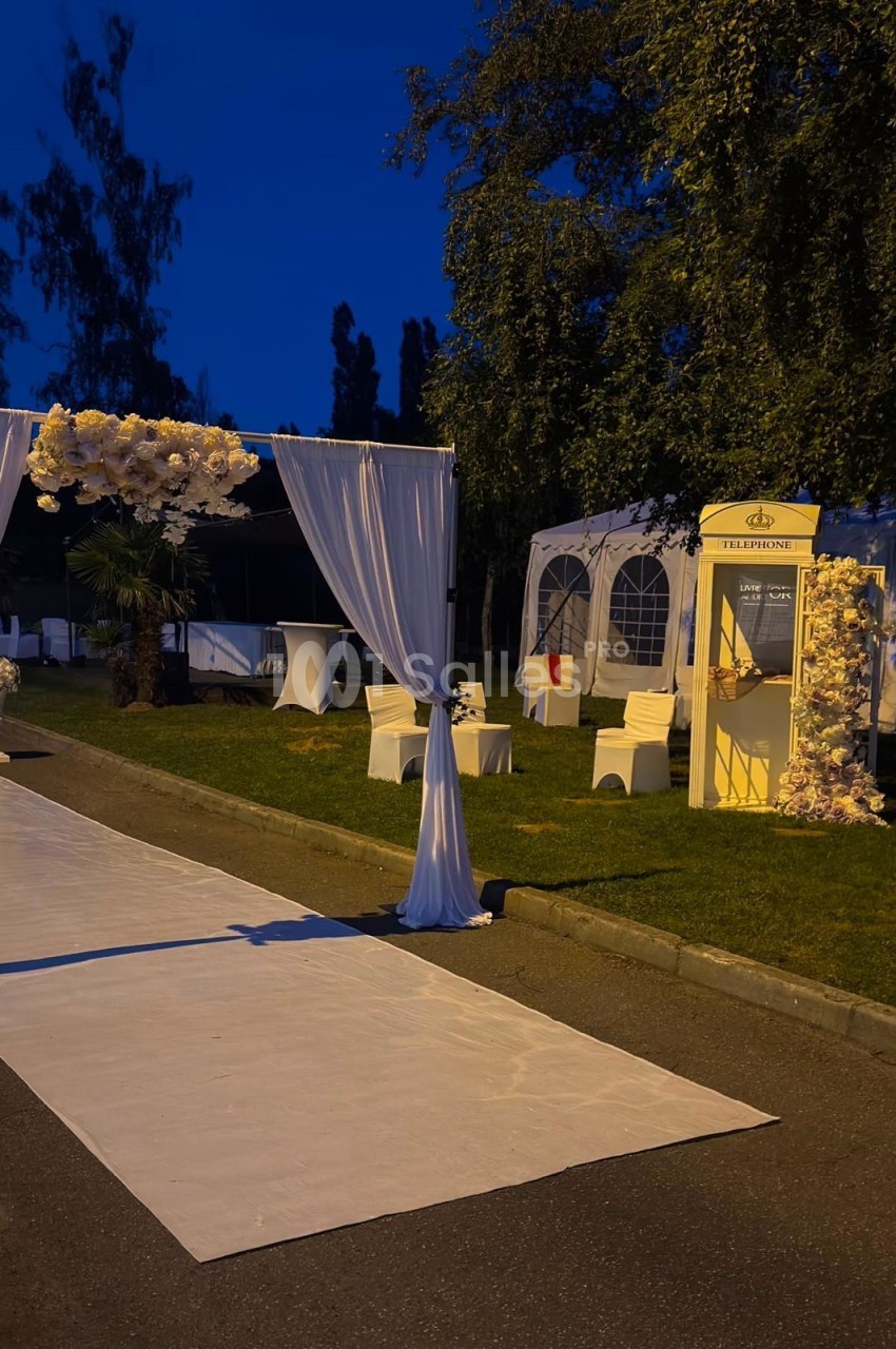 Allée décorée pour un événement en soirée, avec arche florale, tapis blanc et cabine téléphonique ornée de fleurs.