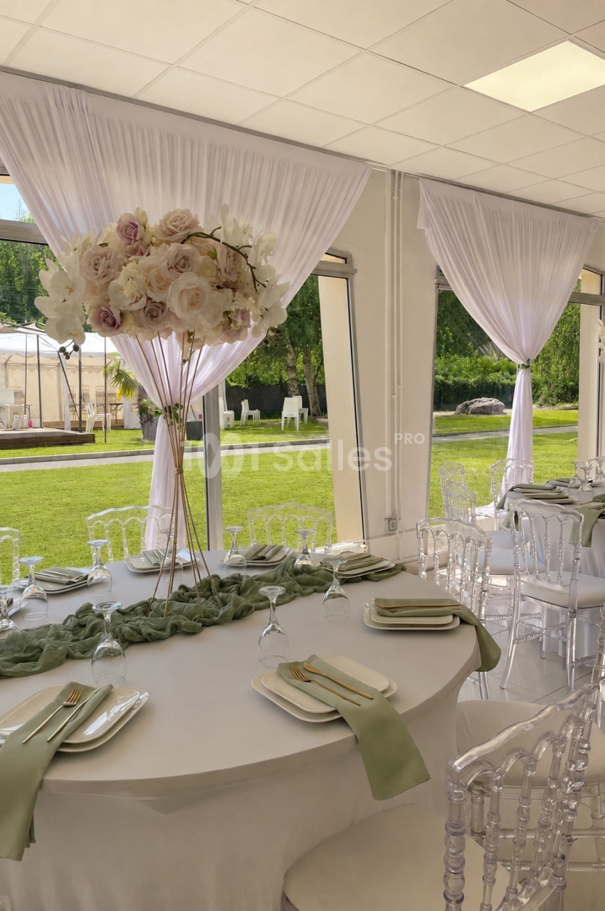 Salle de réception lumineuse avec tables décorées de nappes blanches, serviettes vertes et centres de table fleuris.