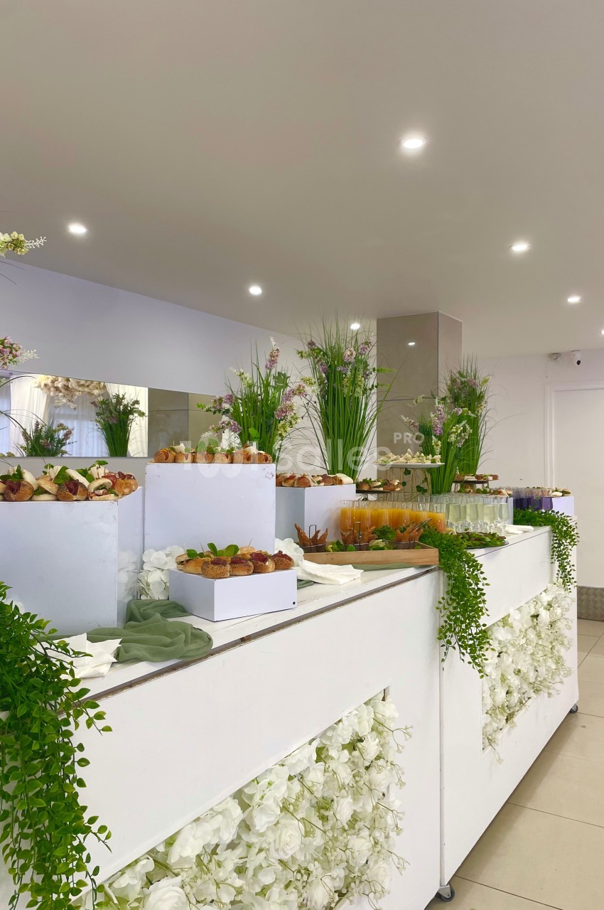 Buffet décoré de fleurs blanches et vertes, avec des plateaux de fruits et de verrines dans une salle lumineuse.