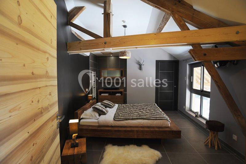 Location salle Vignieu (Isère) - Domaine de Suzel #4 Chambre moderne avec poutres apparentes, lit double, murs sombres, tapis en fourrure et grande fenêtre laissant entrer la…