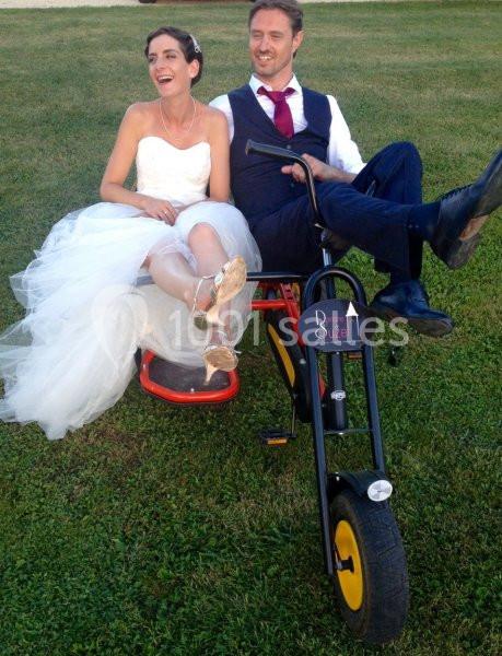 Location salle Vignieu (Isère) - Domaine de Suzel #20 Un couple en tenue de mariage assis sur un tricycle noir dans un jardin.