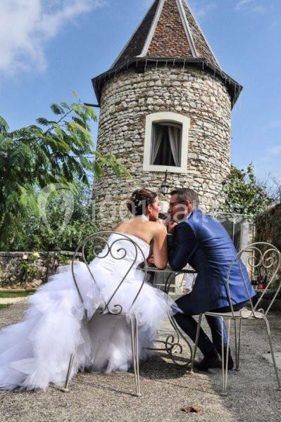 Location salle Vignieu (Isère) - Domaine de Suzel #13 Un couple en tenue de mariage assis à une table en fer forgé devant une petite tour en pierre.