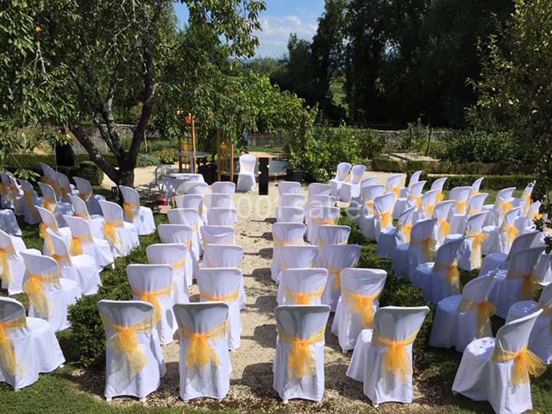 Location salle Vignieu (Isère) - Domaine de Suzel #14 Chaises blanches décorées de nœuds jaunes disposées en rangées dans un jardin pour une cérémonie en plein air.