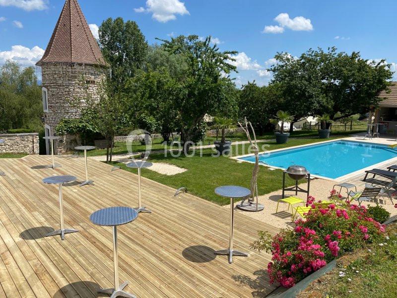 Location salle Vignieu (Isère) - Domaine de Suzel #24 Terrasse en bois avec tables hautes, piscine, jardin arboré et tour en pierre sous un ciel bleu.