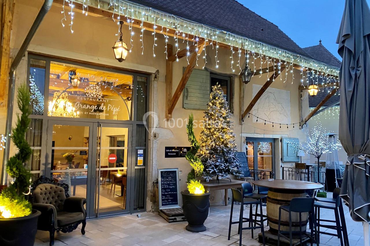Location salle Vignieu (Isère) - Domaine de Suzel #11 Entrée d'un restaurant décorée pour les fêtes avec guirlandes lumineuses, sapin enneigé et mobilier de terrasse.