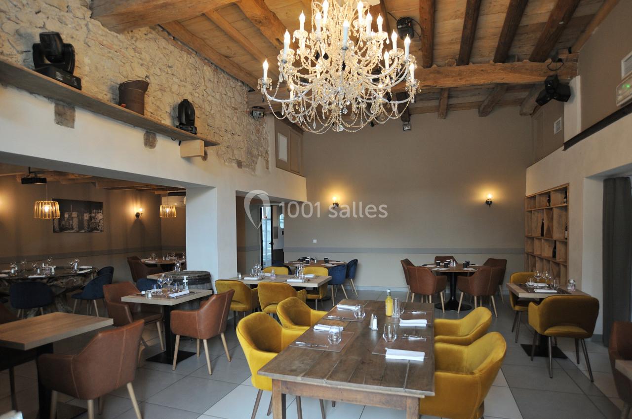 Location salle Vignieu (Isère) - Domaine de Suzel #6 Salle de restaurant lumineuse avec murs en pierre, poutres apparentes, lustre en cristal et mobilier moderne coloré.
