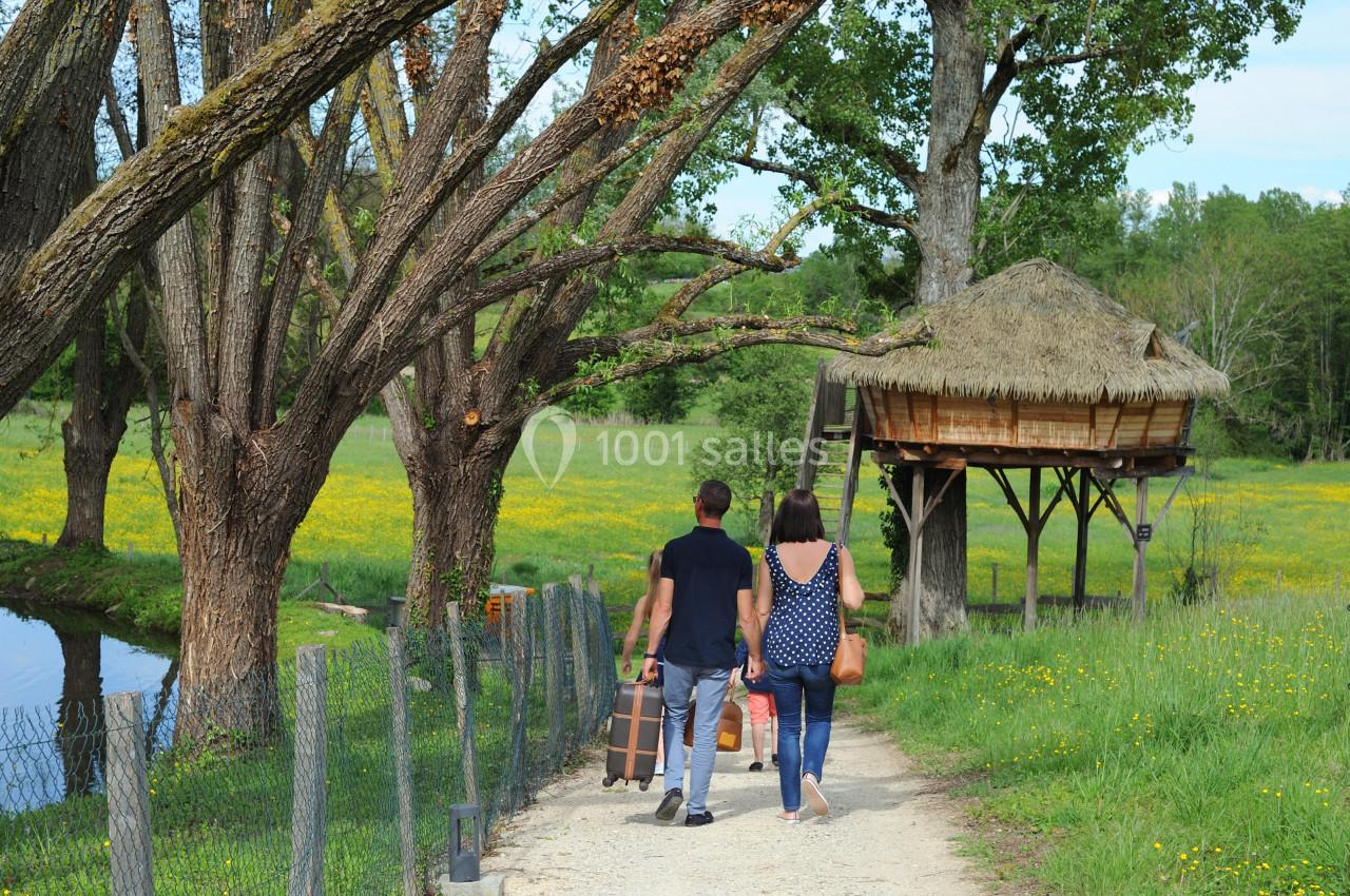 Location salle Vignieu (Isère) - Domaine de Suzel #28 Un couple marche sur un chemin bordé d'arbres, transportant des valises près d'une cabane en bois sur pilotis.
