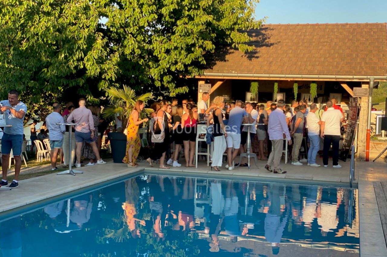 Location salle Vignieu (Isère) - Domaine de Suzel #34 Groupe de personnes rassemblées près d'une piscine, devant un bar extérieur, sous un ciel dégagé en fin de journée.