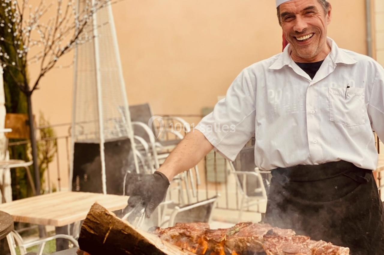 Location salle Vignieu (Isère) - Domaine de Suzel #35 Un cuisinier souriant en tenue blanche prépare des grillades sur un barbecue en extérieur.