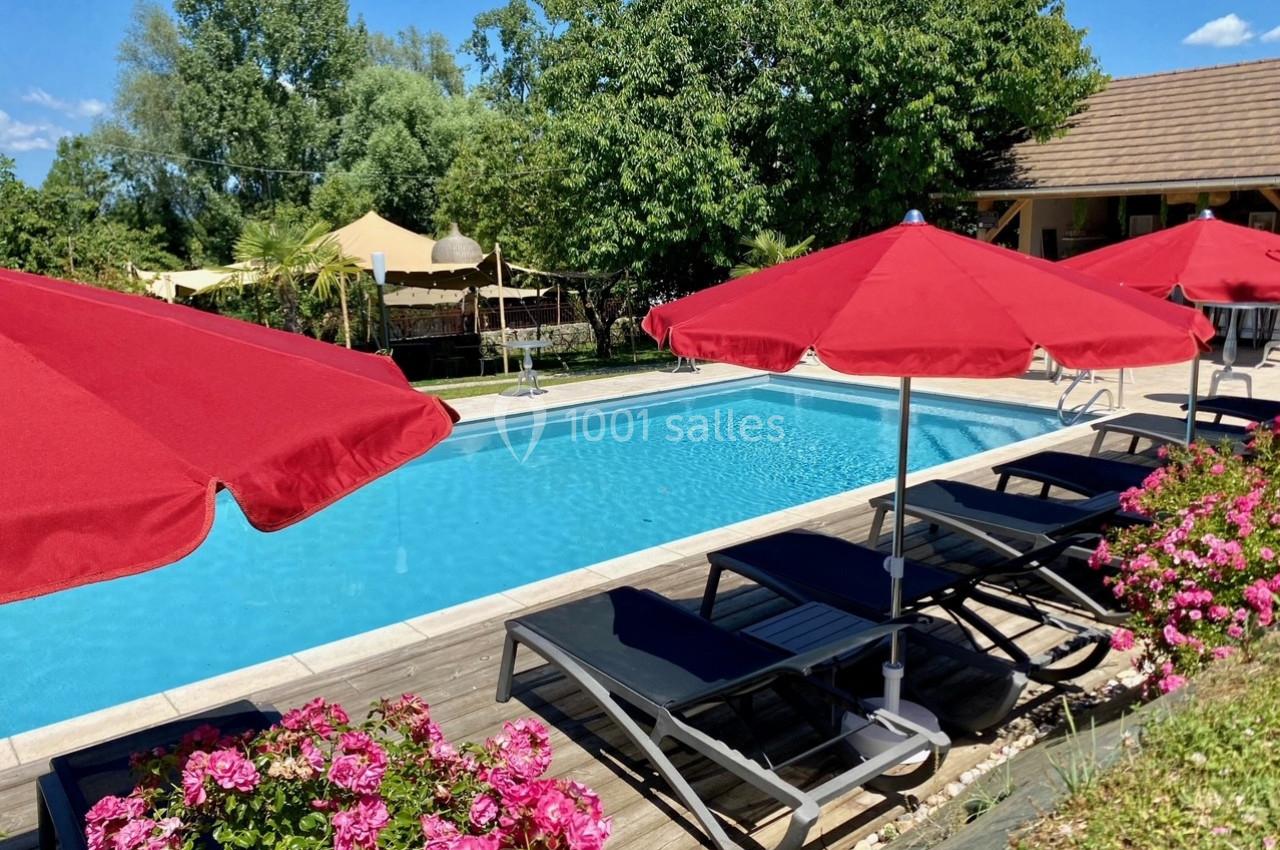 Location salle Vignieu (Isère) - Domaine de Suzel #36 Piscine extérieure entourée de transats noirs, parasols rouges, fleurs roses et végétation verdoyante.
