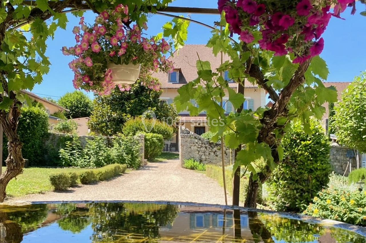 Location salle Vignieu (Isère) - Domaine de Suzel #38 Allée de jardin bordée de verdure et de fleurs suspendues, menant à une maison avec un ciel dégagé en arrière-plan.