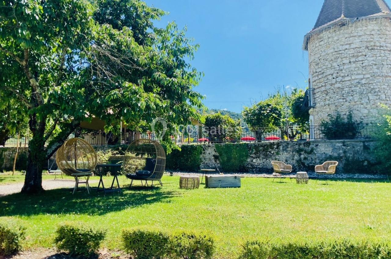 Location salle Vignieu (Isère) - Domaine de Suzel #39 Jardin verdoyant avec chaises en rotin, arbres ombragés et une tour en pierre en arrière-plan.