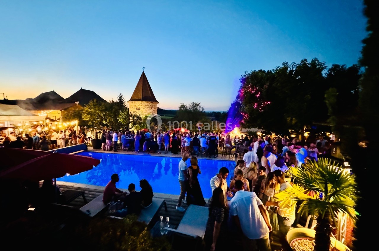 Location salle Vignieu (Isère) - Domaine de Suzel #46 Foule rassemblée autour d'une piscine éclairée en soirée, avec un feu d'artifice en arrière-plan et un bâtiment en pierre.