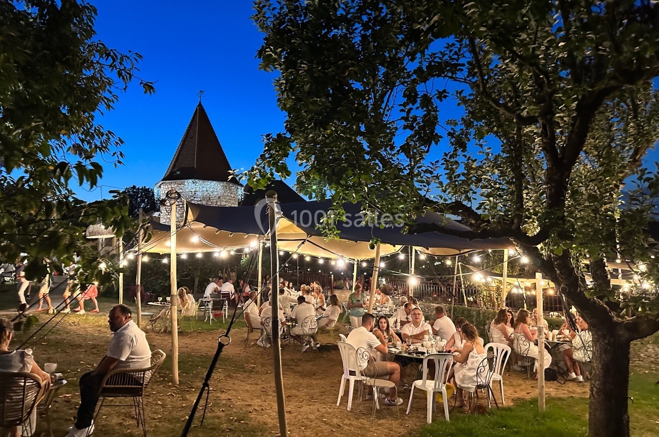 Location salle Vignieu (Isère) - Domaine de Suzel #48 Des personnes dînent en plein air sous une tente éclairée, entourées d'arbres, avec une église en arrière-plan au crépuscule.