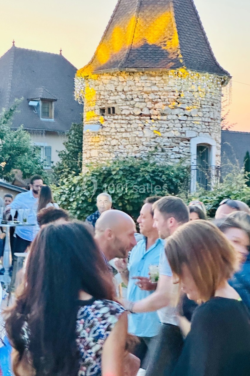 Location salle Vignieu (Isère) - Domaine de Suzel #49 Groupe de personnes discutant lors d'un événement en plein air devant une tour en pierre au coucher du soleil.