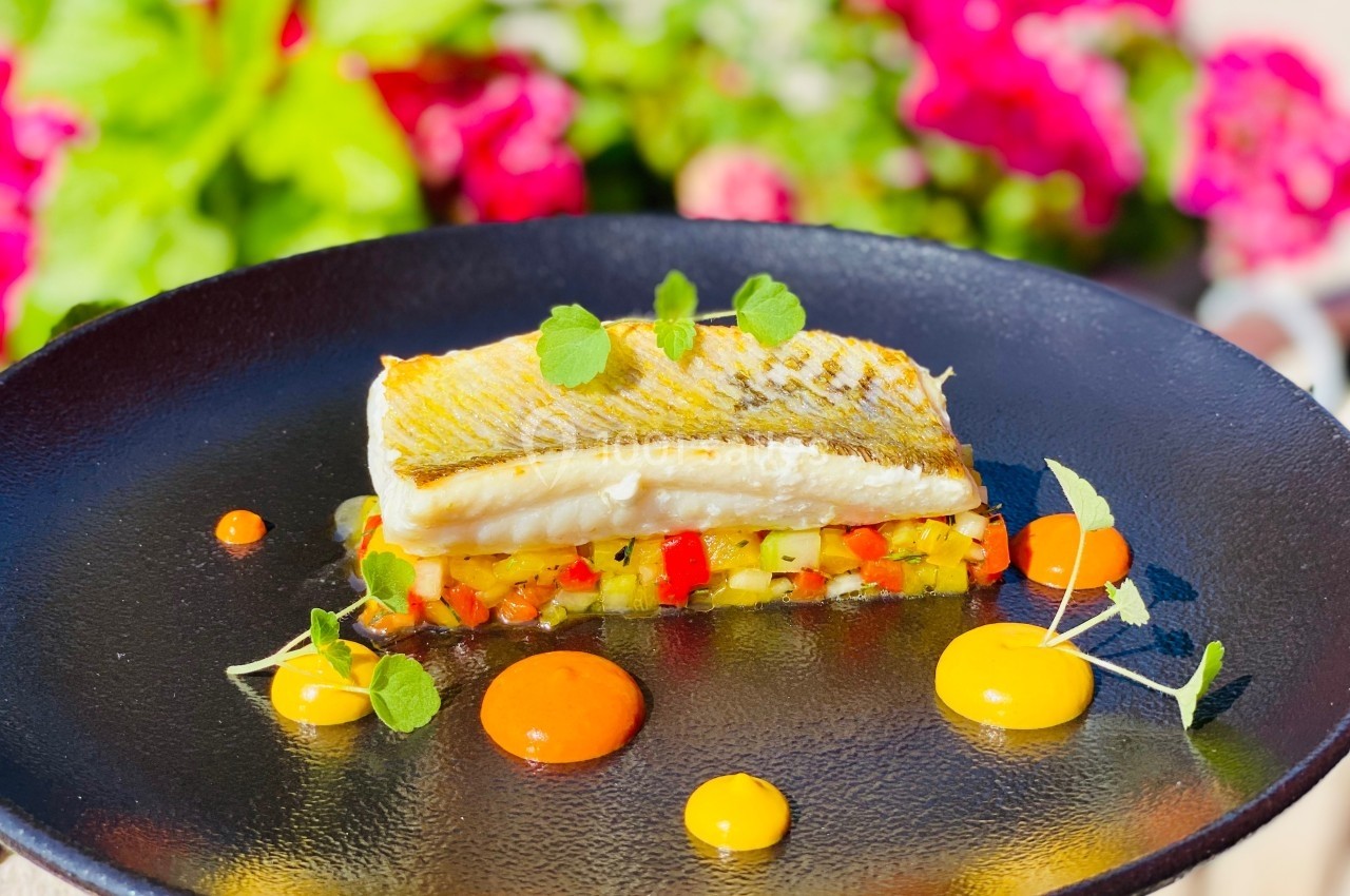 Location salle Vignieu (Isère) - Domaine de Suzel #51 Filet de poisson grillé sur lit de légumes colorés, décoré de sauces et herbes, avec un fond de fleurs floues.