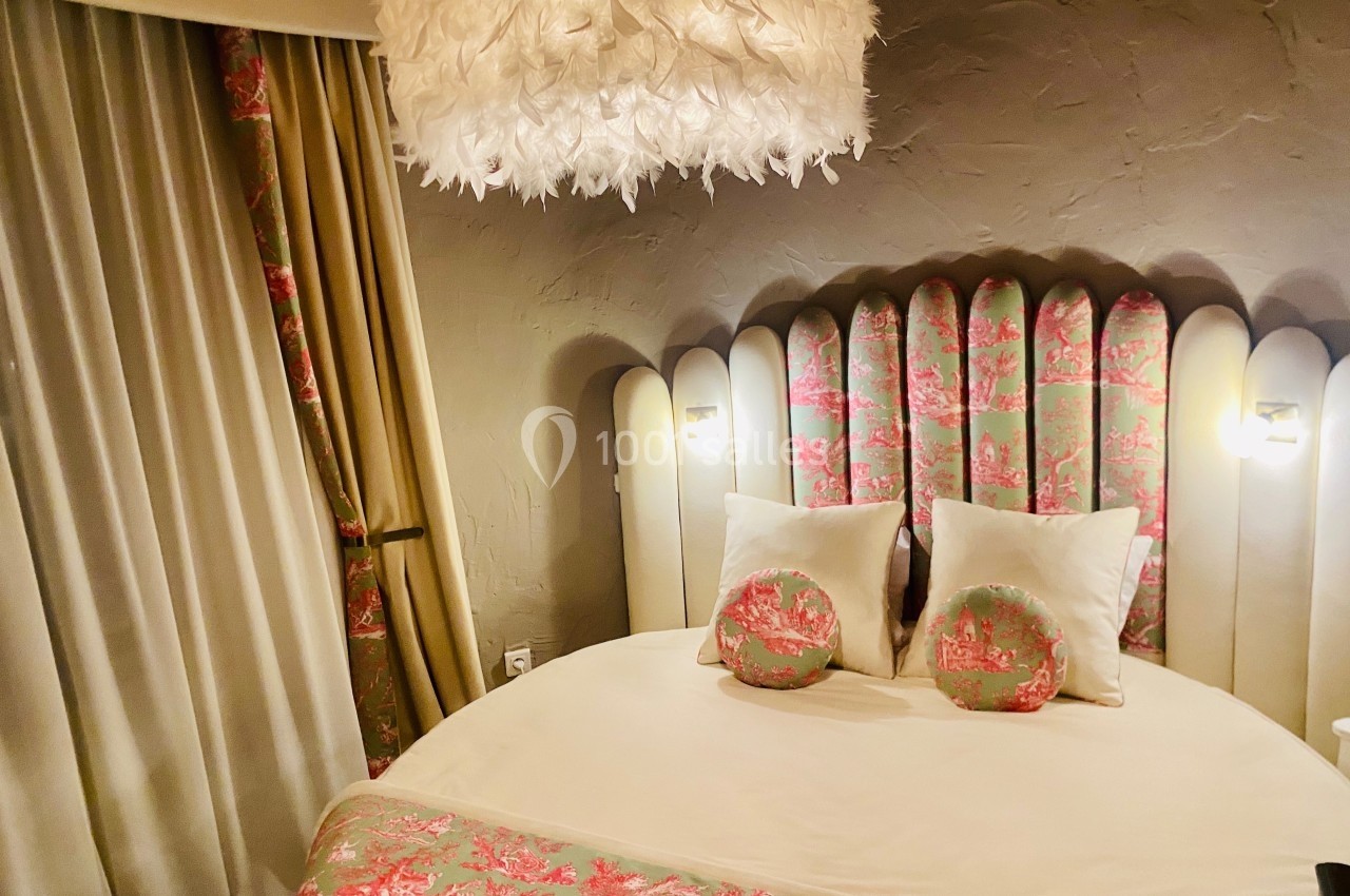 Location salle Vignieu (Isère) - Domaine de Suzel #54 Lit rond avec tête de lit capitonnée à motifs floraux, éclairé par des appliques murales et un lustre à plumes blanches.