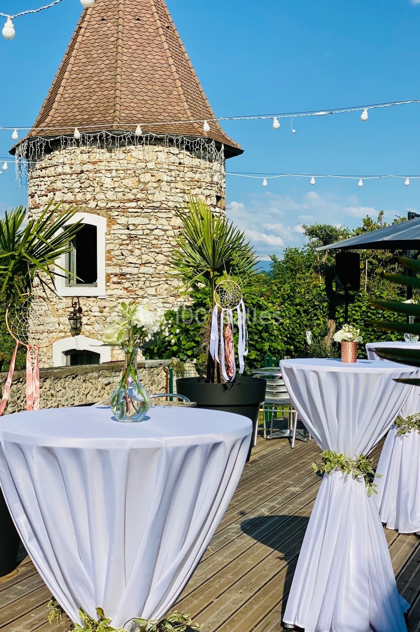 Location salle Vignieu (Isère) - Domaine de Suzel #57 Terrasse ensoleillée avec tables hautes décorées, tour en pierre en arrière-plan et guirlandes lumineuses suspendues.