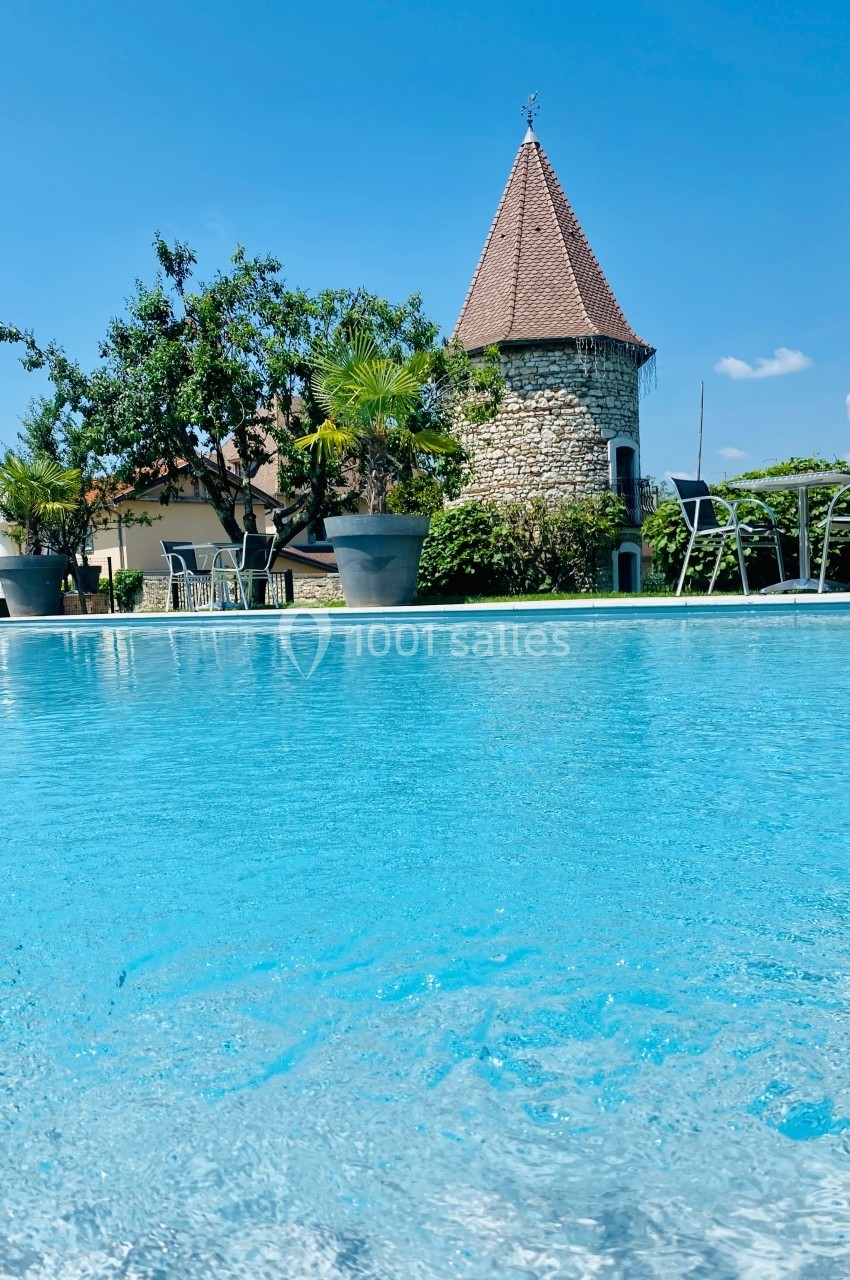 Location salle Vignieu (Isère) - Domaine de Suzel #58 Piscine extérieure avec eau turquoise, entourée de chaises, plantes en pots et un bâtiment en pierre avec toit en pointe.