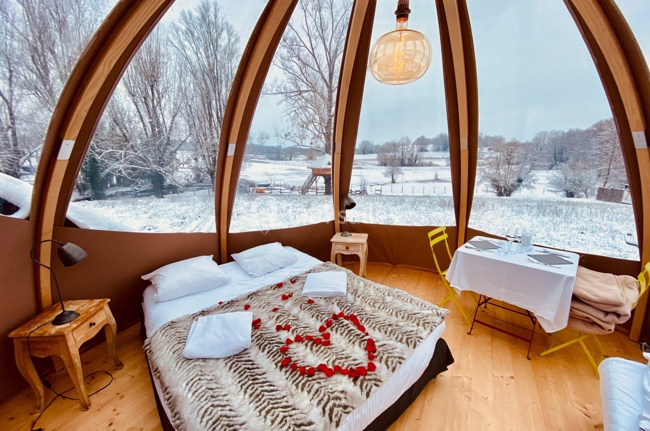 Location salle Vignieu (Isère) - Domaine de Suzel #59 Intérieur d'une bulle en bois avec lit décoré de pétales de rose, vue sur un paysage enneigé à travers de grandes baies…