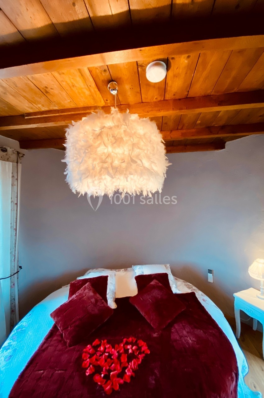 Location salle Vignieu (Isère) - Domaine de Suzel #60 Chambre avec lit rond, couverture rouge, pétales de rose, lampe de chevet et plafond en bois avec suspension plumeuse.