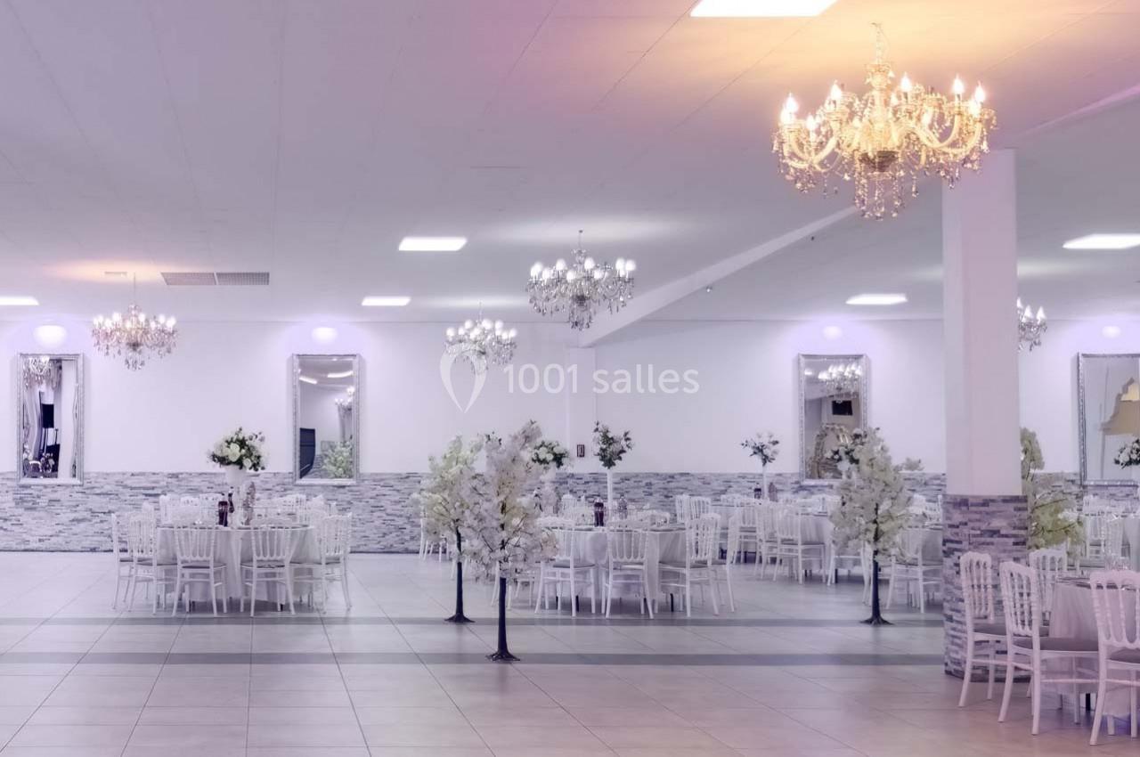 Salle de réception lumineuse avec tables décorées, chaises blanches et lustres élégants suspendus au plafond.