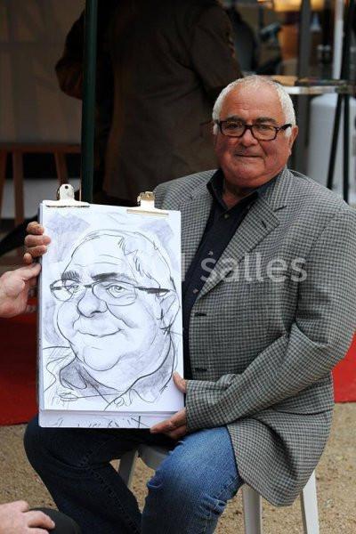 Location salle Vignieu (Isère) - Domaine de Suzel #10 Un homme assis tenant un dessin caricatural de lui-même, réalisé sur une feuille attachée à une planchette.