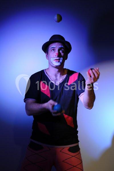 Location salle Vignieu (Isère) - Domaine de Suzel #12 Un jongleur maquillé en lumière bleue jongle avec trois balles, portant un chapeau et des vêtements colorés.