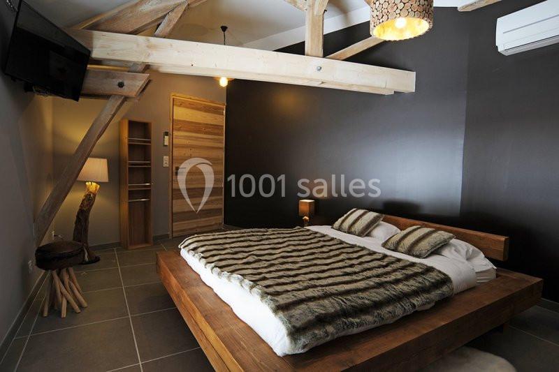 Location salle Vignieu (Isère) - Domaine de Suzel #16 Chambre avec lit double en bois, couverture à motif, poutres apparentes, mur sombre et éclairage tamisé.