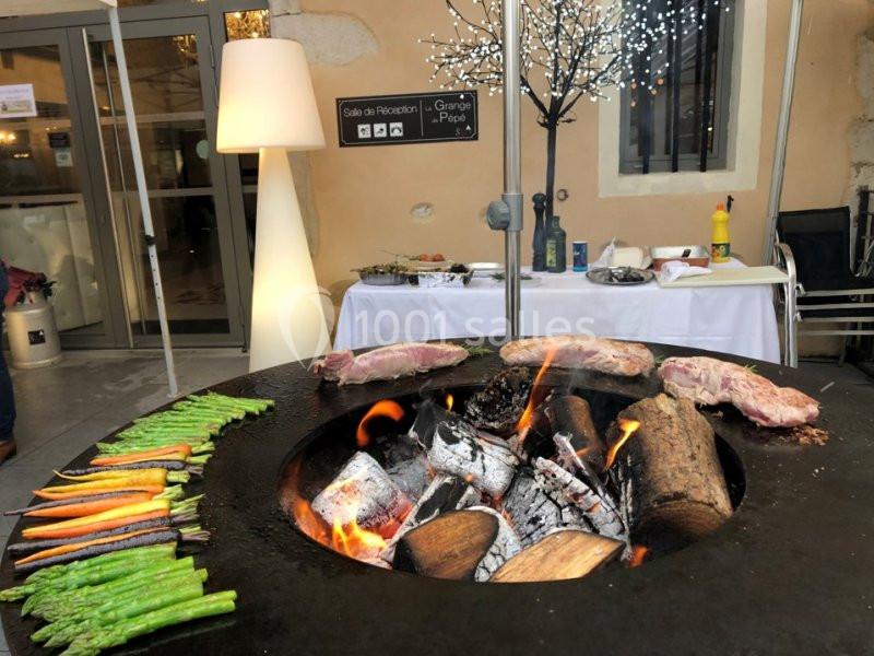 Location salle Vignieu (Isère) - Domaine de Suzel #8 Légumes et viandes grillant sur un barbecue circulaire au feu de bois, avec une table dressée en arrière-plan.