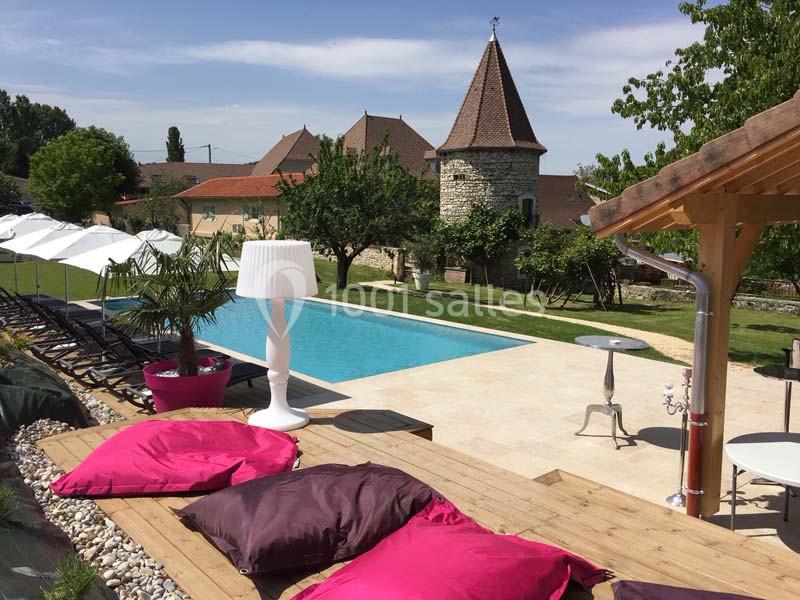 Location salle Vignieu (Isère) - Domaine de Suzel #4 Piscine extérieure entourée de transats, parasols, coussins colorés et vue sur un bâtiment en pierre avec une tour.