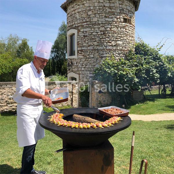 Location salle Vignieu (Isère) - Domaine de Suzel #5 Un chef en tenue blanche cuisine des brochettes et de la viande sur un barbecue rond dans un jardin près d'une tour en…