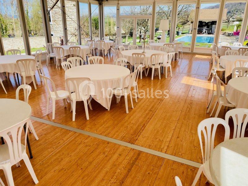 Location salle Vignieu (Isère) - Domaine de Suzel #6 Salle lumineuse avec tables rondes recouvertes de nappes blanches et chaises en plastique, vue sur un jardin et une piscine.