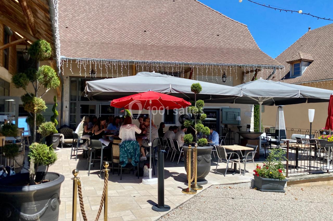Location salle Vignieu (Isère) - Domaine de Suzel #19 Terrasse extérieure d'un restaurant avec des tables occupées, parasols gris et rouge, et décorations végétales.