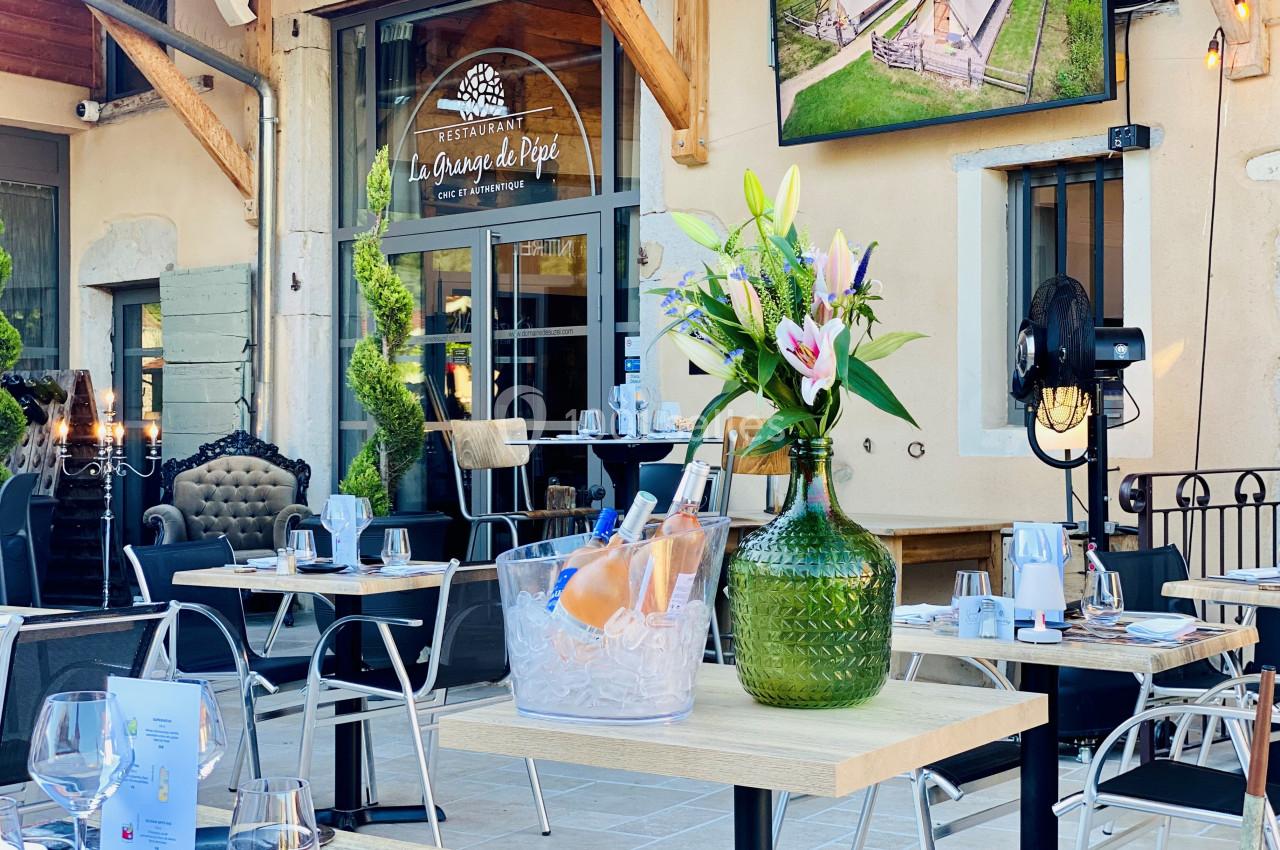 Location salle Vignieu (Isère) - Domaine de Suzel #23 Terrasse d’un restaurant avec tables dressées, vase de fleurs et seau à glace contenant des bouteilles de vin.