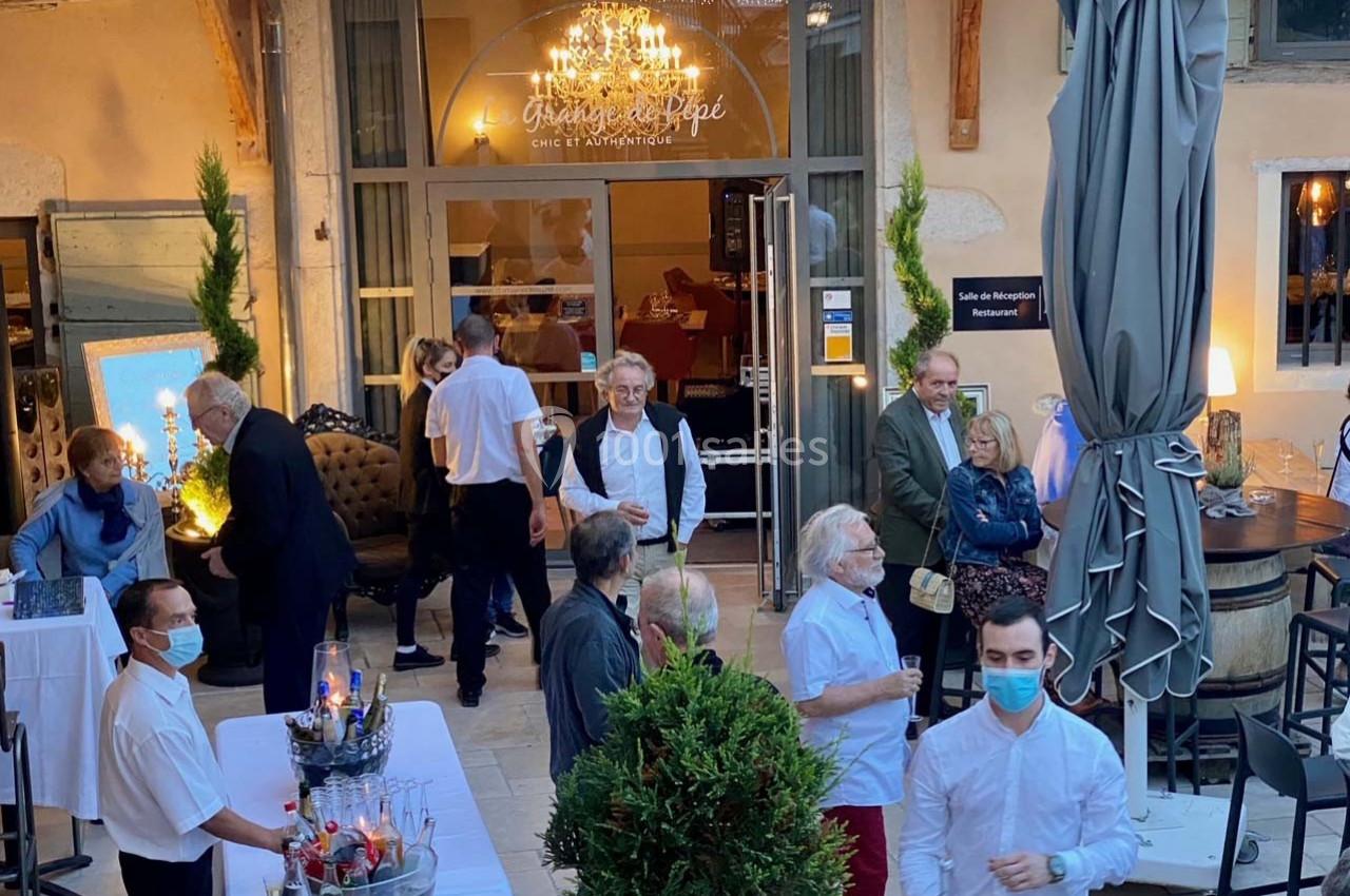 Location salle Vignieu (Isère) - Domaine de Suzel #26 Groupe de personnes discutant dans une cour extérieure aménagée avec tables, plantes et éclairage décoratif.