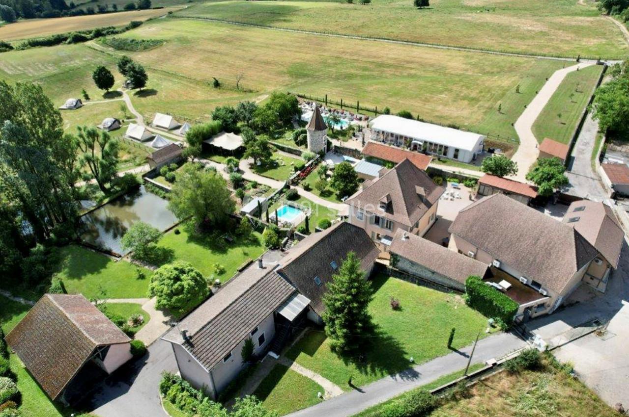 Location salle Vignieu (Isère) - Domaine de Suzel #2 Vue aérienne d'un domaine rural avec plusieurs bâtiments, un étang, une piscine et des espaces verts environnants.
