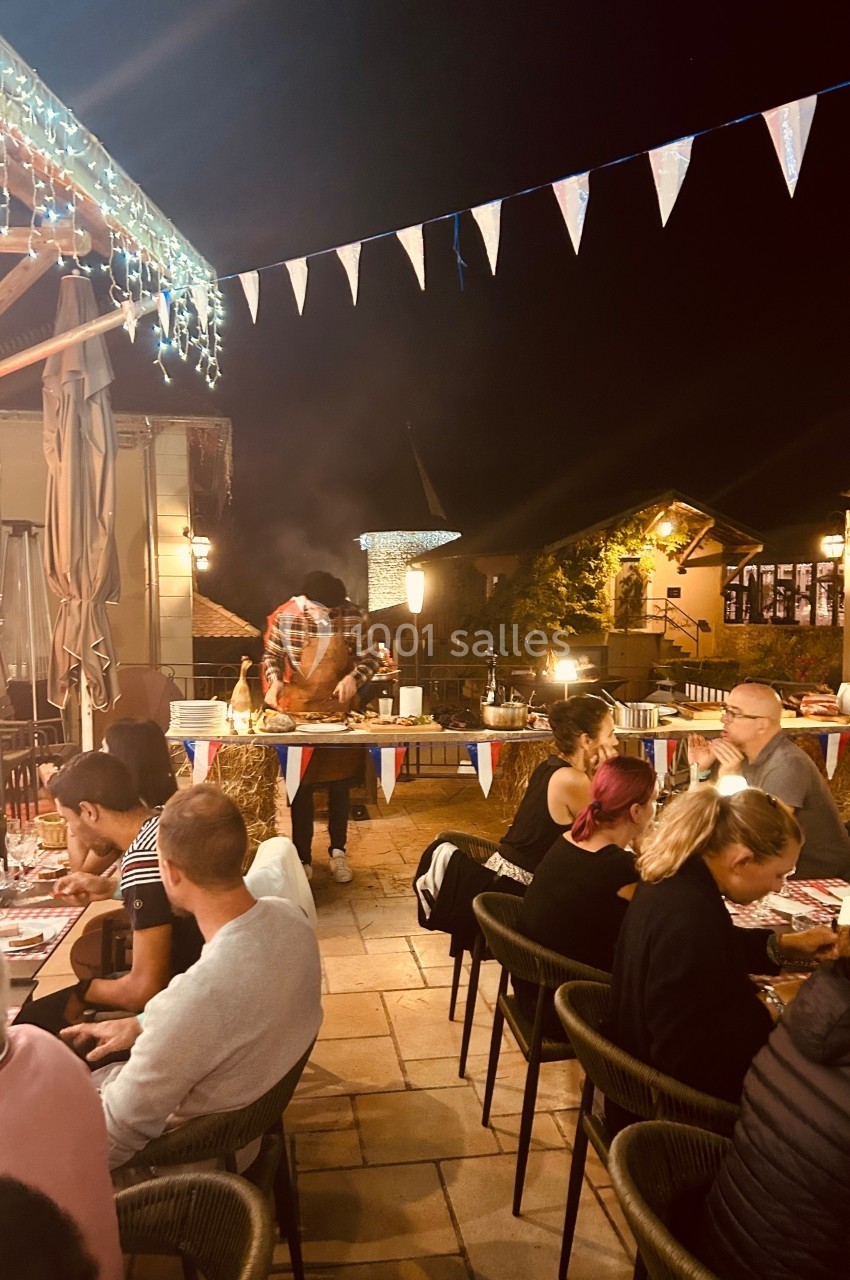 Location salle Vignieu (Isère) - Domaine de Suzel #46 Terrasse animée d'un restaurant en soirée, avec des convives attablés et un cuisinier préparant des plats en extérieur.