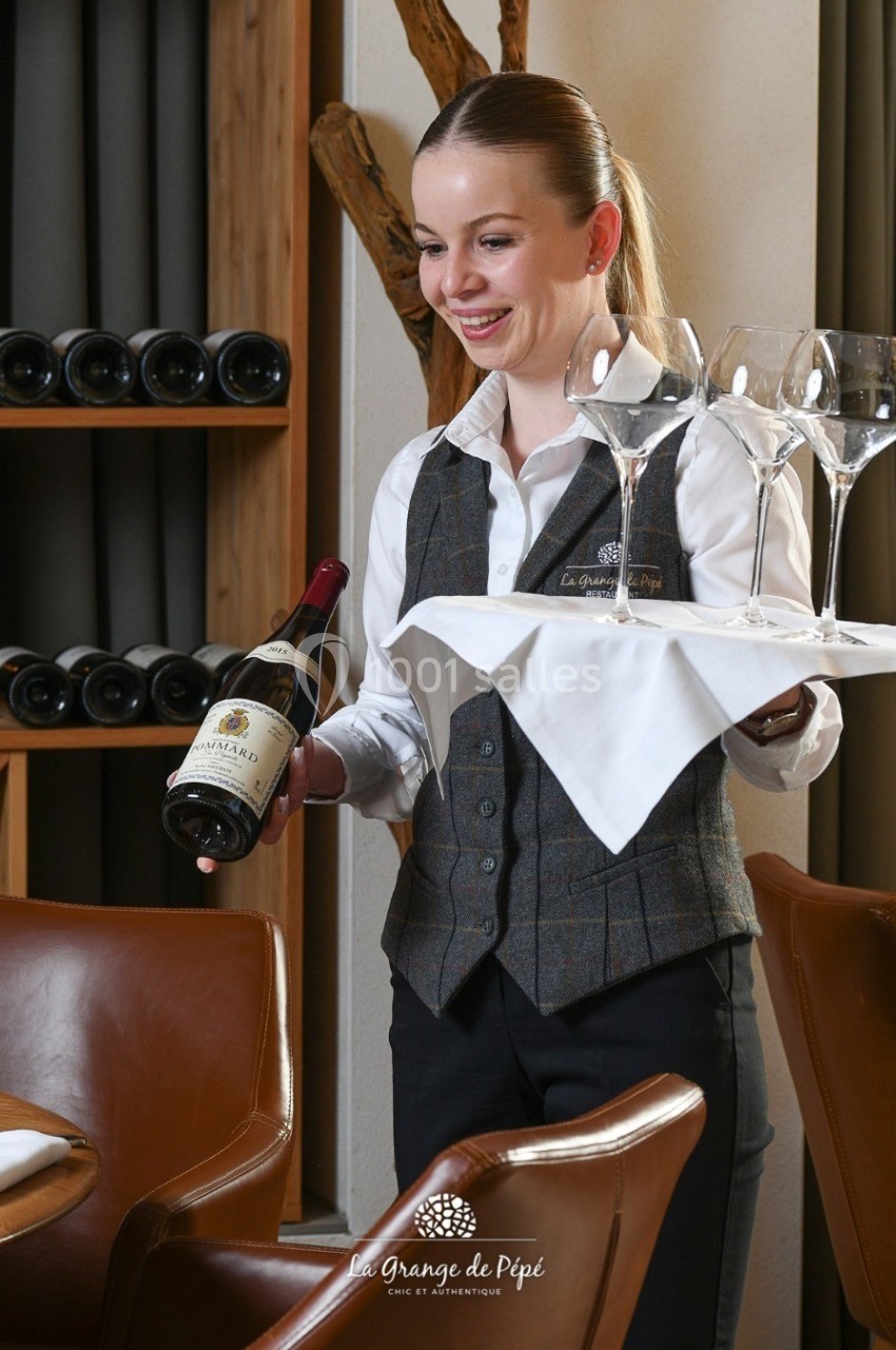 Serveuse souriante tenant un plateau avec des verres et une bouteille de vin dans un restaurant élégant.
