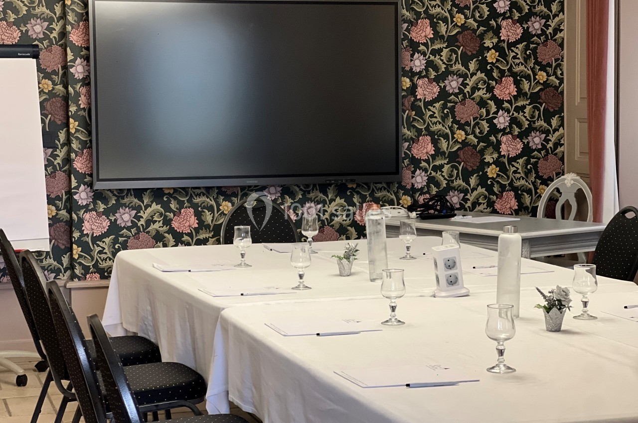Salle de réunion avec table rectangulaire dressée, chaises noires, écran mural et fond de papier peint fleuri.