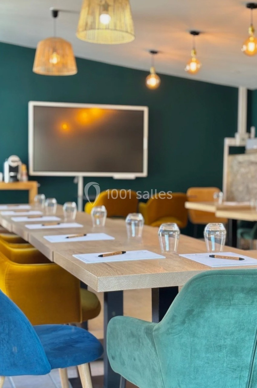 Salle de réunion moderne avec table en bois, chaises colorées, écran, et éclairage suspendu.