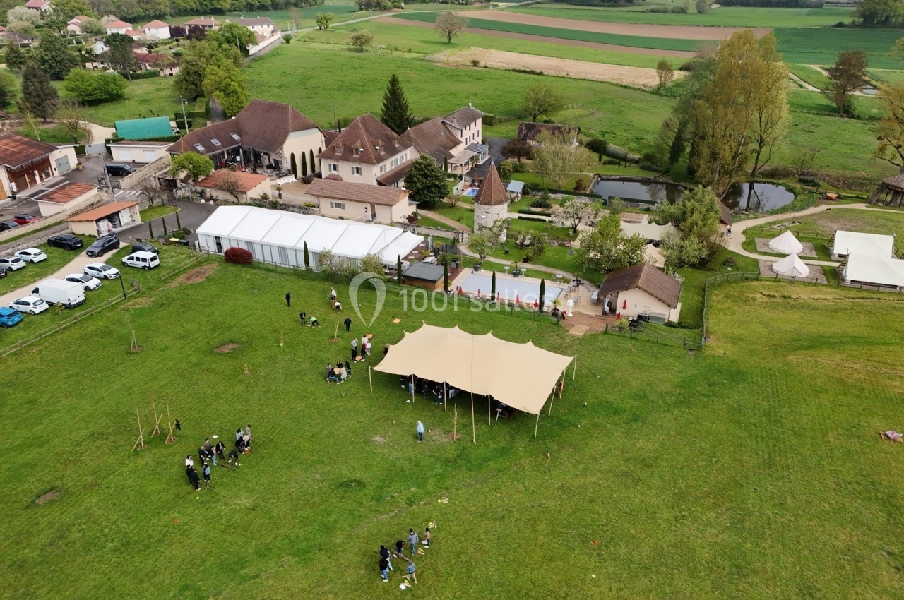 Vue aérienne d'un domaine rural avec des bâtiments, une tente beige, des espaces verts et des participants dispersés.