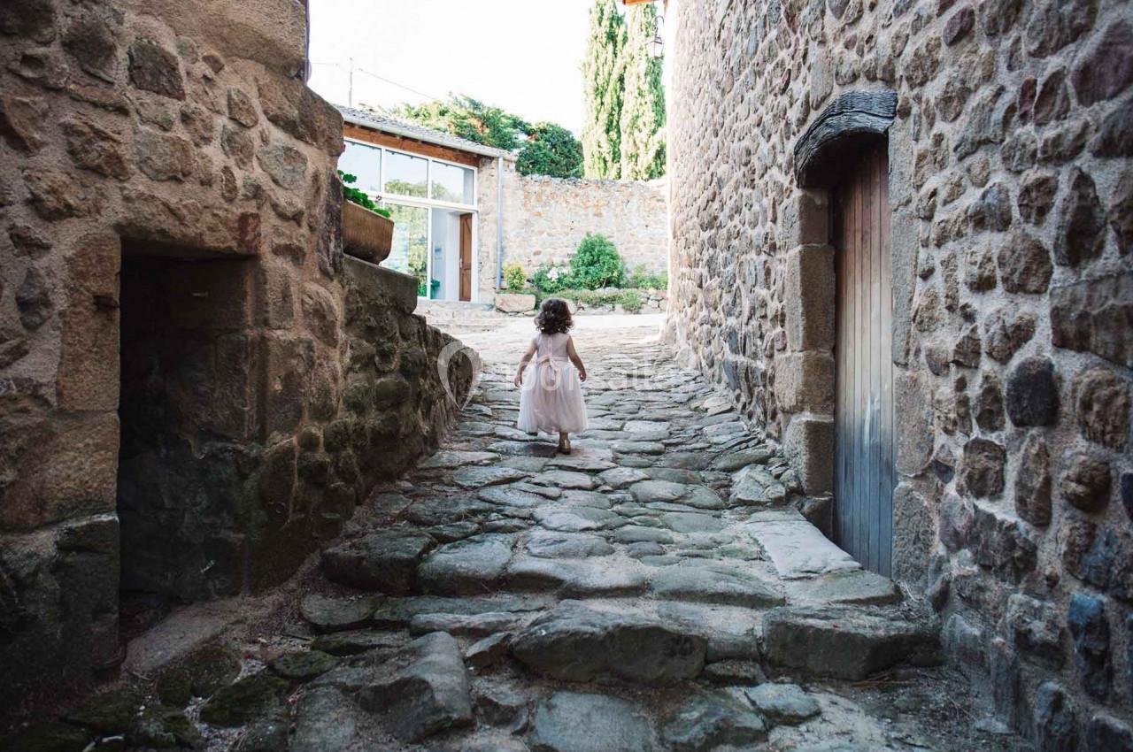 Une petite fille en robe marche sur un chemin pavé entre des bâtiments en pierre dans un village calme.