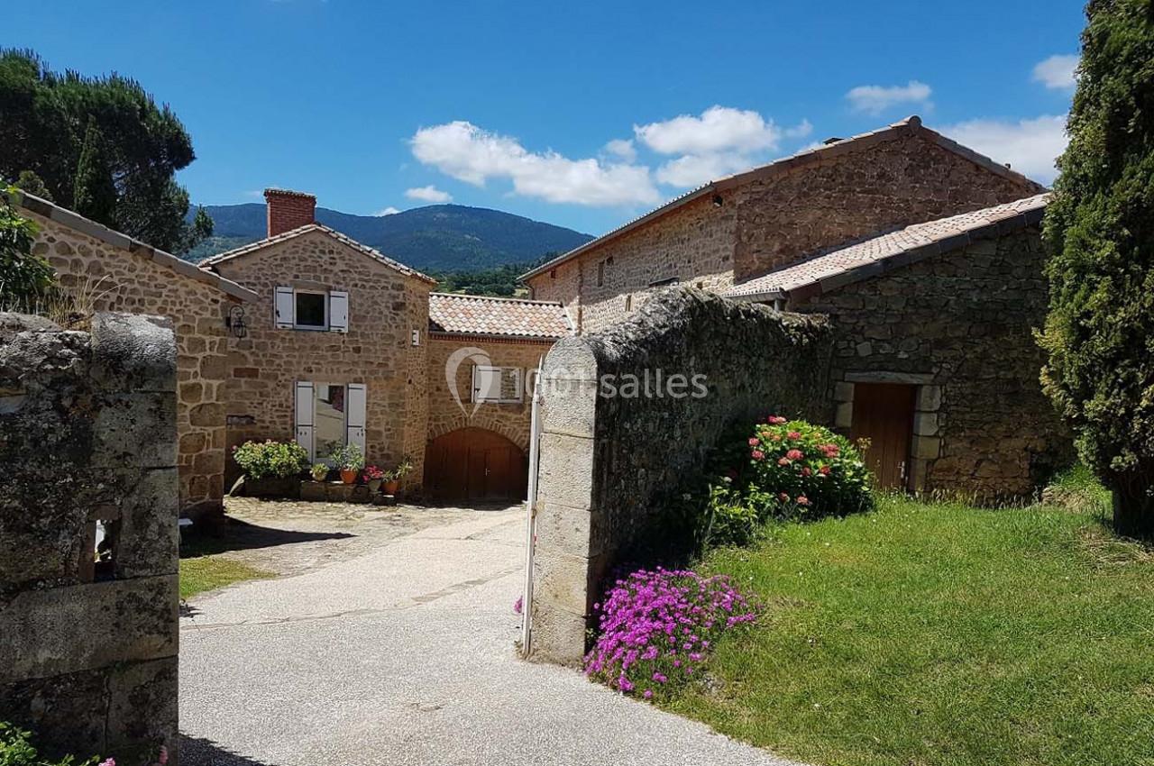Cour d'une maison en pierre avec fleurs colorées, entourée de bâtiments rustiques et vue sur des collines boisées.