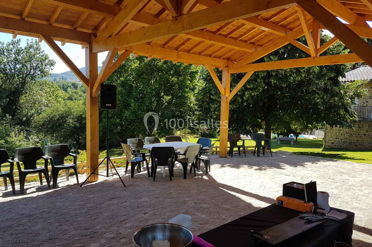 Structure en bois avec tables et chaises disposées sur un sol en gravier, entourée de verdure et d'arbres.