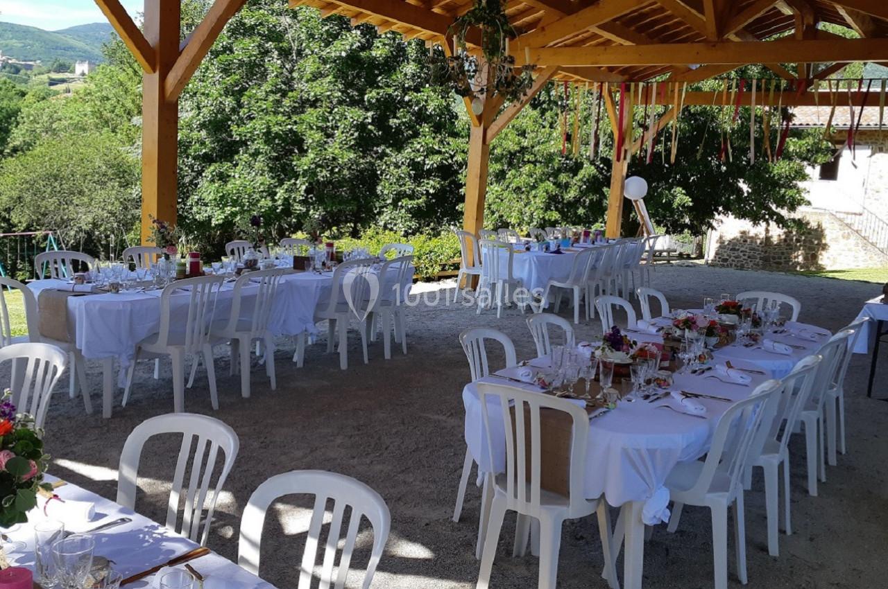Tables dressées pour un repas en plein air sous une pergola en bois, entourée de verdure.