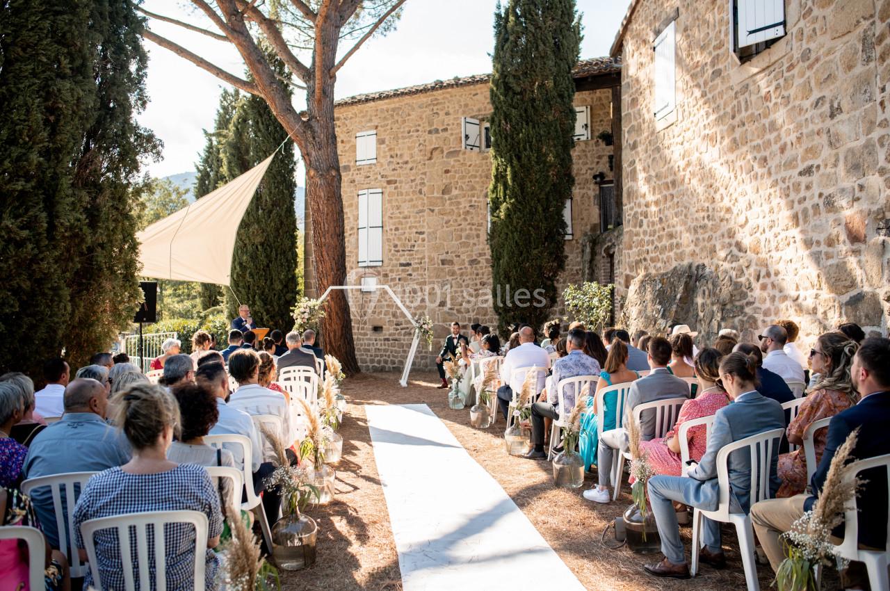 Cérémonie de mariage en plein air avec des invités assis, devant un bâtiment en pierre entouré de cyprès.